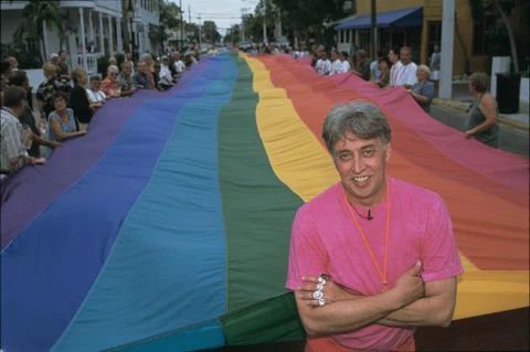 Gilbert Baker and the Birth of the Rainbow Flag

In this historically rich photograph, activist and artist Gilbert Baker stands proudly in front of an enormous rainbow flag—the original symbol he designed in 1978 that would become an enduring emblem of LGBTQ+ pride around the world. Dressed in a bright pink shirt, arms confidently crossed, Baker smiles as the massive flag unfurls behind him, held up by a long line of supporters. The image is both personal and political, capturing the joy, defiance, and artistry that Baker poured into his creation. The backdrop of a city street lined with parade-goers underscores the impact of Baker’s work, as what was once a grassroots design stitched by hand now stretches across entire blocks, waved by people who may not even know the name of its creator—but carry his legacy nonetheless.

The rainbow flag was not just a graphic statement; it was born out of urgent necessity. After the assassination of gay rights icon Harvey Milk and the rising backlash against queer communities, Baker sought a symbol that would unify and uplift. Inspired by movements of civil rights and social liberation, and steeped in his background as an artist and drag performer, he saw the rainbow as a spectrum that could include everyone. The first version had eight stripes, each with specific meanings: pink for sex, red for life, orange for healing, yellow for sunlight, green for nature, turquoise for magic, indigo for serenity, and violet for spirit. As production evolved, it was pared down to the six-color version more commonly used today, but its symbolic depth remained intact.

This photograph, though casual in composition, captures a watershed moment in cultural history. Baker’s smile radiates fulfillment, but the scale of the flag behind him reminds us of the collective effort it took to bring the LGBTQ+ movement into public consciousness. It’s a quiet portrait of revolution—one stitched in fabric, carried by many, and still rippling through time.