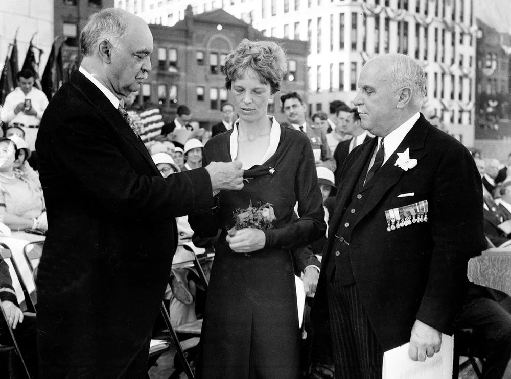 Honoring a Legend – Amelia Earhart Receives the Distinguished Flying Cross : 
A Milestone in Aviation and Gender Equality

This evocative historical image captures Amelia Earhart standing with quiet poise as she receives the Distinguished Flying Cross on June 24, 1932—a moment that cemented her status as a pioneering force in aviation. Dressed modestly and holding a bouquet of flowers, Earhart is flanked by officials and onlookers as a dignitary pins the medal to her chest. This scene commemorates her record-setting solo transatlantic flight and marks the first time a woman received the prestigious honor, awarded by the U.S. Congress for extraordinary achievement in aerial flight. Just a month earlier, on May 20–21, 1932, Earhart had flown alone across the Atlantic from Newfoundland to Northern Ireland in 14 hours and 56 minutes, piloting a Lockheed Vega 5B. She became not only the first woman but only the second person in history to complete such a journey, following Charles Lindbergh.

The crowd surrounding her reflects both awe and respect—a mix of military figures, civic leaders, and members of the public gathered to witness the recognition of a groundbreaking accomplishment. Earhart’s calm, introspective expression seems to transcend the moment, as if she’s already thinking ahead to future adventures. The medal ceremony, though brief, stands as a cultural landmark, symbolizing not only personal courage and technical skill but also the broader evolution of women’s roles in society. This image memorializes a point in time when one woman’s flight helped shift public perception and inspired generations to aim higher—both literally and figuratively.