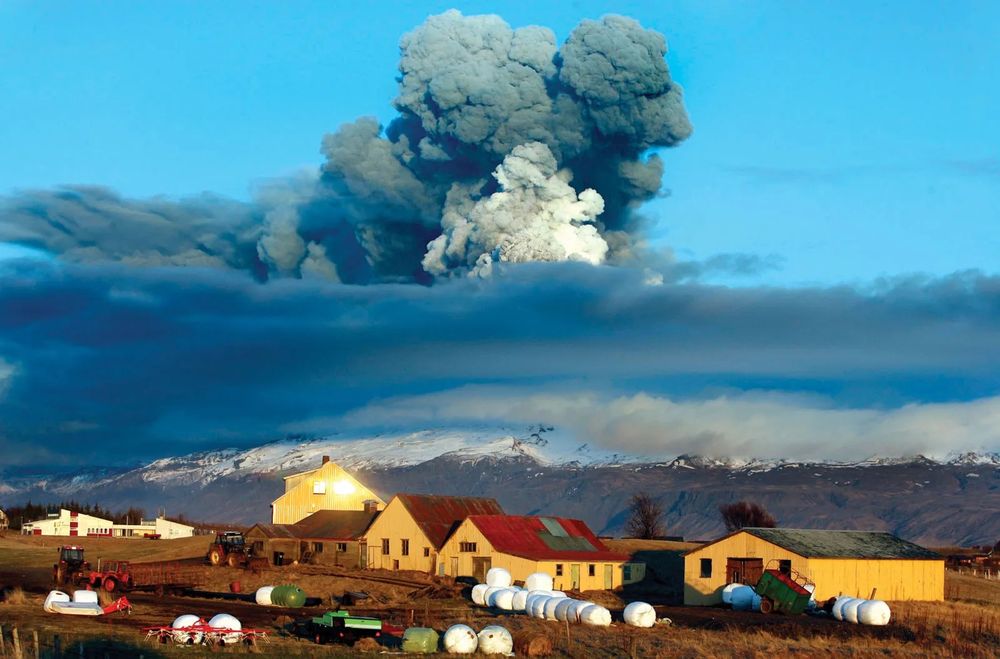 Eyjafjallajökull Ash Cloud Over Icelandic Farmland – A Modern Disruption Rooted in Ancient Power

This image captures the distant ash plume of the Eyjafjallajökull volcano rising ominously above a pastoral Icelandic farm on a crisp, clear day. Taken during the 2010 eruption that began in April and officially ceased on May 22, the photo shows an expansive volcanic cloud contrasting dramatically with the quaint, scattered yellow and red-roofed buildings in the foreground. Wrapped in early morning light, the farm structures and snow-dusted mountains provide a peaceful yet surreal setting for one of the 21st century’s most disruptive natural events. The eruption not only released massive quantities of ash and volcanic gas into the upper atmosphere, but also paralyzed much of Europe’s air travel. Over 100,000 flights were canceled in the span of a week, marking the largest air traffic shutdown since World War II. The photo’s visual balance between human development and natural upheaval encapsulates the geographic and cultural realities of living in Iceland, where volcanism is not just a geological phenomenon, but a formative element of national identity. The vibrant cloud formations in the sky—including darker ash and lighter water vapor—illustrate how quickly conditions can shift, affecting agriculture, health, and communication systems. The eruption’s impact rippled beyond Iceland, prompting discussions on aviation safety, volcanic forecasting, and the fragile interdependence of global networks. This photograph is not just a snapshot of natural drama—it is a lens into the vulnerability of modern systems when confronted with Earth’s untamed forces, and a tribute to the resilience of Icelandic communities long accustomed to coexisting with fire and ice.