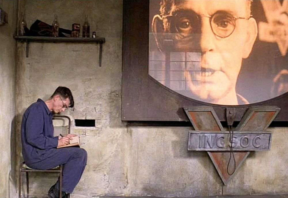 Surveillance and Subversion – A Scene from the 1984 Film Adaptation of Nineteen Eighty-Four

This image captures a hauntingly accurate visual interpretation of George Orwell’s dystopian world as depicted in the 1984 film adaptation of Nineteen Eighty-Four, directed by Michael Radford. The frame centers on Winston Smith, the novel’s protagonist, seated alone in a dilapidated, gray-washed room that bears the stark and worn texture of authoritarian decay. He is hunched over a notebook, quietly committing to paper the rebellious act of thought—an action fraught with danger in the regime of Big Brother. The dim lighting and confined space convey a sense of both isolation and suffocation, echoing the oppressive psychological atmosphere of Oceania.

Dominating the right half of the image is a massive telescreen projecting the ominous visage of Big Brother, whose eyes appear to follow the viewer, reinforcing the regime’s ceaseless surveillance. The screen is mounted above the symbol of INGSOC, the Party’s ideology, shaped like a downward-pointing V flanked by authoritarian steel and a taut cord—a visual metaphor for control and punishment. The televised face speaks not only to the propaganda constantly broadcast to the citizens, but also to the inescapable gaze that defines the narrative’s central conflict.

This visual sequence encapsulates the novel’s most chilling themes: the erosion of privacy, the control of truth, and the existential weight of living under constant observation. It also emphasizes the loneliness of dissent in a world engineered to erase it. Winston’s small act of writing represents the last vestige of human autonomy in a society where language has been sanitized and emotion criminalized.

The scene resonates deeply with contemporary viewers, especially in an age of digital surveillance, data collection, and algorithmic manipulation.