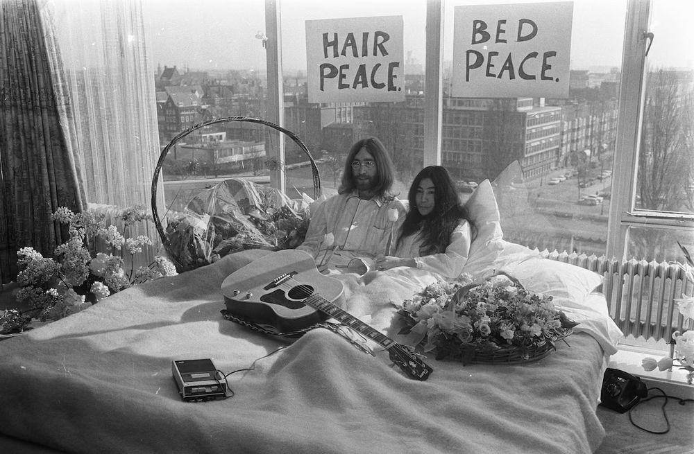 Lennon and Ono’s Montreal Bed-In Begins (1969)

This black-and-white image captures the beginning of John Lennon and Yoko Ono’s second “Bed-In for Peace,” launched on May 26, 1969, at the Queen Elizabeth Hotel in Montreal. Sitting upright in a large hotel bed, Lennon and Ono are dressed in white pajamas, surrounded by flowers, a cassette recorder, and an acoustic guitar — the very one later used to record the antiwar anthem “Give Peace a Chance.” Large, hand-drawn signs behind them declare “HAIR PEACE” and “BED PEACE,” intentionally simple messages that would become globally recognized symbols of their protest. The window behind them reveals the Montreal cityscape, a quiet contrast to the political noise they hoped to disrupt. Unlike conventional protests, the Bed-In was meant to be peaceful, absurd, and unignorable — leveraging Lennon’s fame to draw cameras and conversations into a conversation about peace during the Vietnam War era. The media frenzy it sparked brought journalists, activists, and artists into their room across several days, turning a hotel suite into an unexpected epicenter of global activism. Their Montreal Bed-In followed a similar event in Amsterdam earlier that year and was conceived as a response to escalating conflict, not just in Vietnam, but globally. This iconic image freezes a moment when pop culture and pacifism met with intentionality — using celebrity as a megaphone for peace. The recording of “Give Peace a Chance” would follow days later in this very room, with dozens of guests joining the chorus, immortalizing this unconventional protest in both music and memory.