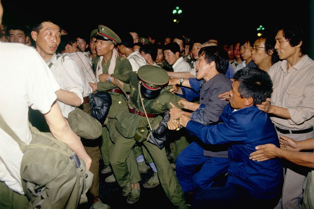 Clashes Between Protesters and Soldiers During the Crackdown

This intense image captures a chaotic and emotional scene from the night of June 3 into the early hours of June 4, 1989, during the violent crackdown on the Tiananmen Square protests. A group of civilians is seen struggling with uniformed Chinese soldiers, some trying to hold them back while others shout, restrain, or confront them face-to-face. The soldiers, wearing olive-green military uniforms and red-collared caps, carry gear and are visibly overwhelmed by the resistance they’re facing. Some are mid-action, pulling away or being shoved, their expressions tense and bewildered. One soldier is bent at the waist, his face obscured, caught in the push-and-pull of the fray. The civilians—mostly middle-aged men and younger activists—grapple with them, faces contorted in anguish, fear, or determination. This confrontation is part of the larger government operation to retake Tiananmen Square by force, as the Chinese Communist Party leadership declared martial law to quell six weeks of growing public dissent. Although tanks and armored vehicles are more often associated with this moment, these close-range skirmishes between unarmed citizens and heavily equipped troops were frequent throughout Beijing that night. The streetlights in the background barely illuminate the crowd, lending a claustrophobic and urgent energy to the image. The photograph reflects the desperation and bravery of ordinary citizens willing to resist military might to protect the spirit of peaceful protest. These nighttime confrontations left an unknown number dead and wounded, as the Chinese government has never released an official death toll. This moment serves as a stark visual reminder of the human cost of authoritarian control and the enduring power of civilian resistance.