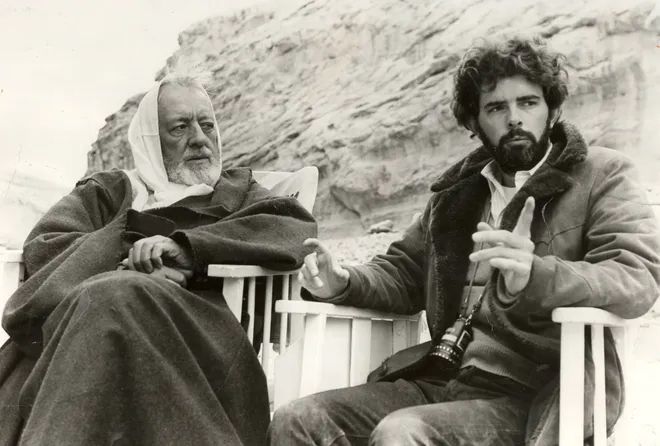 Vision and Legacy – George Lucas Directs Alec Guinness on the Set of Star Wars

This behind-the-scenes black-and-white photograph captures a pivotal moment in cinematic history: George Lucas directing the esteemed Alec Guinness during the filming of Star Wars (1977), later known as Episode IV: A New Hope. Seated outdoors amid the rocky terrain of Tunisia—doubling for the desert planet Tatooine—the image radiates a quiet intensity and mutual respect. Lucas, young and bearded with a camera slung around his neck, gestures with intent as he discusses a scene, his hands illustrating a point with precision. His puffy jacket and thoughtful expression reflect the pressures of shaping a bold vision with limited resources and massive ambition. Opposite him, Guinness—already a legendary British actor known for roles in The Bridge on the River Kwai and Lawrence of Arabia—sits in Obi-Wan Kenobi’s Jedi robes, hood up against the wind, listening with focus. Guinness’s participation lent gravitas to the film and anchored its spiritual dimension. His portrayal of Kenobi would become iconic, despite his initial skepticism about the film’s script and science fiction premise. This image encapsulates the unlikely fusion of classical acting tradition and daring new storytelling that Star Wars achieved. It also highlights Lucas’s role not just as a world-builder, but as a careful director balancing legacy talent with a young, unknown cast. This collaboration bridged generations and genres, blending mythic narrative with futuristic setting. The stark desert backdrop and contemplative mood of the shot foreshadow the quiet wisdom and epic conflict embedded in the galaxy-spanning saga that would follow, changing cinema forever.