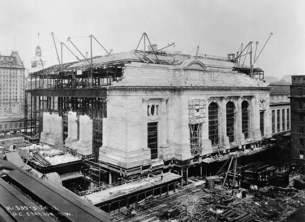 The Construction of Grand Central Terminal (1905-1913)

This black-and-white historical photograph captures Grand Central Terminal in New York City during its construction phase, showcasing its Beaux-Arts architectural style. The grand building, still skeletal, is surrounded by scaffolding, cranes, and construction materials, demonstrating the ambitious scale of the project. The image highlights the transformation of transportation infrastructure in the early 20th century, as the station was designed to accommodate the growing demand for rail travel and electrification. Prior to the terminal’s construction, the site was home to Grand Central Depot (opened in 1871) and later Grand Central Station (1899), which was partially demolished to make way for this vastly expanded structure. The new terminal was designed by the firms Reed & Stem and Warren & Wetmore, featuring a monumental façade, large arched windows, and intricate detailing that exemplified the grandeur of the City Beautiful movement. The construction process required an immense workforce, as workers carved out vast underground spaces for train platforms and waiting areas. Grand Central Terminal was built with modern engineering innovations, including electrified tracks, a two-level concourse system to separate incoming and outgoing trains, and an extensive network of tunnels that would redefine how people traveled through the city. This image encapsulates the meticulous efforts of builders and architects who shaped one of the most iconic train stations in the world. Upon completion in 1913, the station would become the largest in the world, revolutionizing passenger rail service. The construction of Grand Central was part of a broader wave of urban modernization, symbolizing the rise of New York as a global metropolis and serving as a gateway to the nation’s expanding railway network.