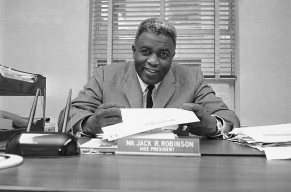 Jackie Robinson the Businessman–Breaking Barriers Beyond the Ballpark

This photo captures Jackie Robinson in a setting few associate with his legacy—behind a desk, in a suit, as “Mr. Jack R. Robinson, Vice President.” Seated with confidence and surrounded by papers, Robinson looks up with a poised expression that merges warmth with professionalism. His nameplate sits front and center, not just as an identifier but as a declaration: the man who shattered baseball’s color barrier was also forging new paths in the world of business

After retiring from baseball in 1956, Robinson refused to be a passive symbol of progress. He became a vocal and active leader in the civil rights movement, using his platform and intellect to push for economic justice. This photo was taken during his tenure as Vice President of Chock full o’Nuts, making him one of the first African Americans to hold a high-level executive position in a major American corporation. It was a radical sight in 1960s America—an era when corporate leadership was overwhelmingly white and segregated in opportunity. Robinson wasn’t there as a figurehead; he was known for attending board meetings, shaping policies, and advocating for better hiring practices and racial equity

The image signifies a powerful evolution: from athlete to activist to corporate trailblazer. Robinson understood that systemic change required more than symbolic victories—it demanded a seat at the table where power and policy were shaped. He didn’t just challenge injustice on the field—he tackled it in boardrooms, banks, and political arenas

The setting and composed demeanor reflect the new phase of Robinson’s mission: economic empowerment for Black Americans. His post-baseball career included founding Freedom National Bank in Harlem and supporting Black-owned businesses. In every role, he insisted on dignity, excellence, and change

Jackie Robinson wasn’t just the man who changed the game—he changed the country, one courageous move at a time