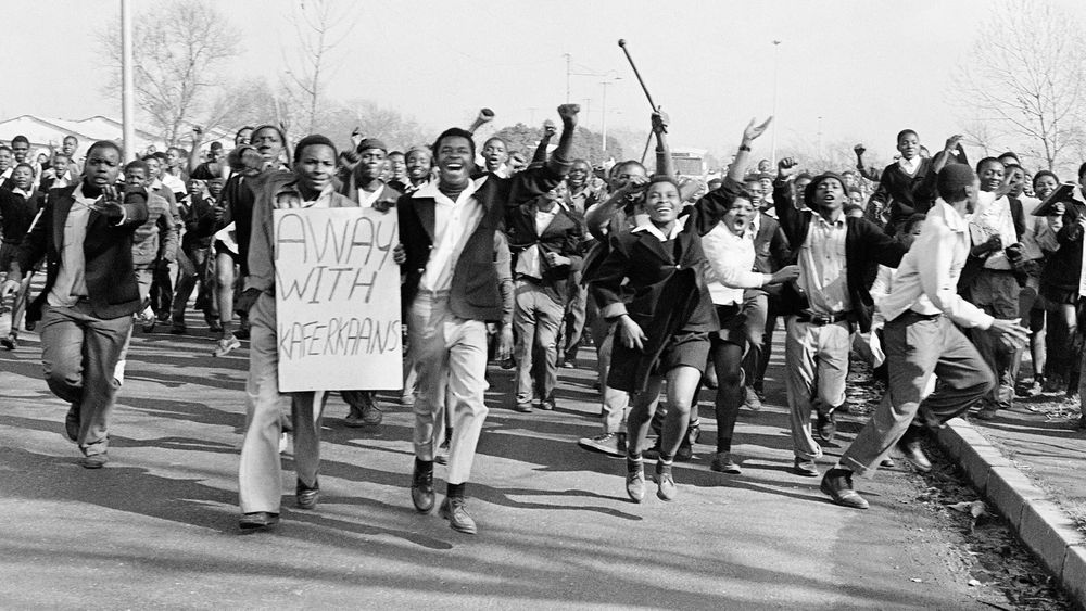 “Away with Afrikaans” – Youth Resistance During the Soweto Uprising

In this powerful black-and-white photo, a large crowd of South African students marches forward with determination and urgency during the Soweto Uprising of 1976. At the forefront, a young man holds a handmade sign reading “AWAY WITH KAAFERKAANS,” a direct protest against the apartheid government’s policy enforcing Afrikaans as a medium of instruction in schools. The faces of the students are filled with resolve, many raising clenched fists—a global symbol of resistance. They wear school uniforms, linking their protest to the educational system they are resisting. The movement is dynamic; the students are in mid-motion, some shouting, others gesturing, their unity palpable as they occupy the street. Behind them, a sea of young protestors stretches into the distance, underscoring the widespread outrage felt across Soweto and beyond. The image captures the early days of what would become a pivotal anti-apartheid movement, sparked on June 16 and continuing through the weeks that followed, including June 20. This image reflects the historical significance of youth activism, the brutality of state oppression, and the symbolic rejection of cultural erasure. The photo has become one of the most iconic visual records of defiance during apartheid, encapsulating the courage and conviction of a generation that challenged one of the most oppressive systems of the 20th century. Their stand laid the moral groundwork for international condemnation of apartheid and galvanized solidarity movements around the globe.