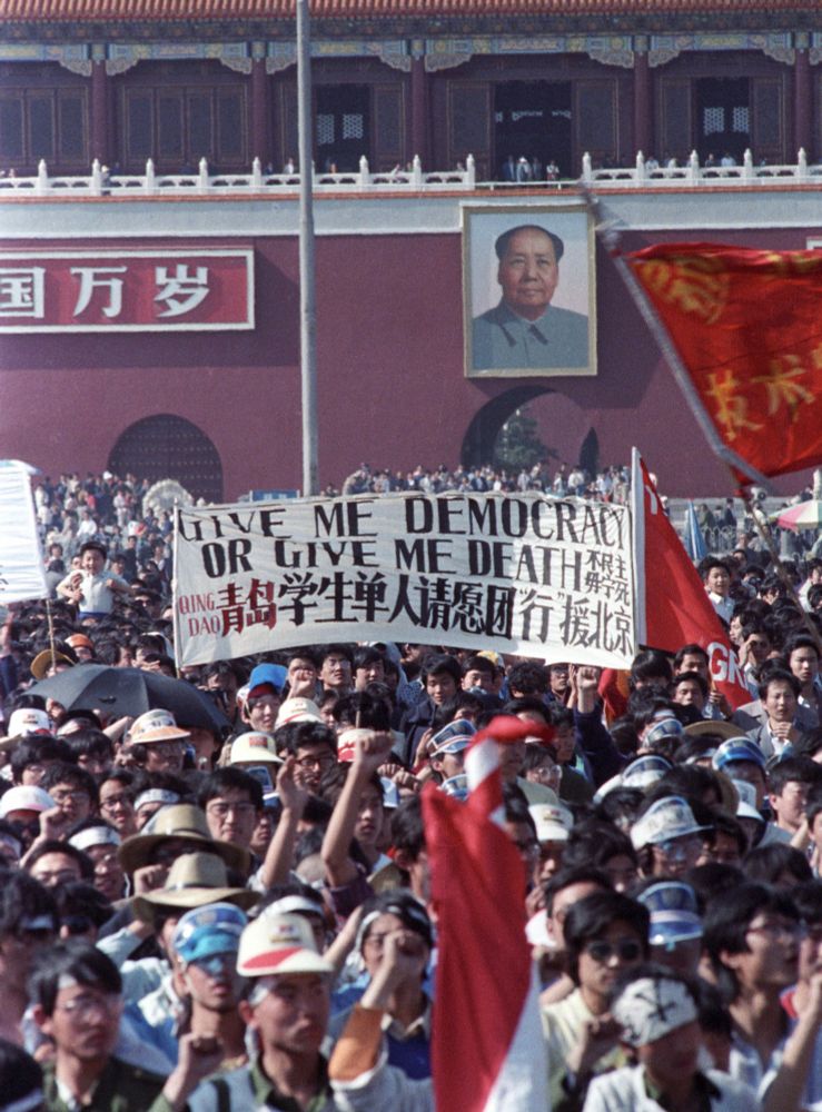 Protest Banner Demanding Democracy in Front of Mao’s Portrait

This striking photograph captures a defining moment during the 1989 Tiananmen Square protests, as thousands of demonstrators stand beneath a banner reading, “Give Me Democracy or Give Me Death.” Positioned directly in front of the Tiananmen Gate and the towering portrait of Mao Zedong—the founder of the People’s Republic of China—the banner juxtaposes the protesters’ urgent call for reform with the enduring symbol of Chinese Communist authority. The crowd, densely packed and unified in purpose, is composed largely of university students, many wearing hats, armbands, or headbands indicating their affiliations with various institutions or activist groups. The sea of raised fists, open mouths mid-chant, and hand-drawn signs reflect a mix of youthful passion and political resolve. At the time, the protest movement was spurred by dissatisfaction with inflation, nepotism, and a lack of political openness following the death of reformist leader Hu Yaobang. What began as a student vigil evolved into a mass demonstration representing a cross-section of Chinese civil society. The emotional and ideological potency of the sign—echoing revolutionary slogans from both East and West—illustrates how deeply the protesters believed in the urgency of their demands. Visible in the background, Mao’s looming visage acts as both a reminder of past authoritarian rule and a symbol of the ideological monolith they sought to challenge. This image encapsulates the extraordinary courage of the protesters and their audacious efforts to claim political agency in a highly controlled system. Today, it remains one of the most iconic visual artifacts from a movement that dared to imagine a different future for China.