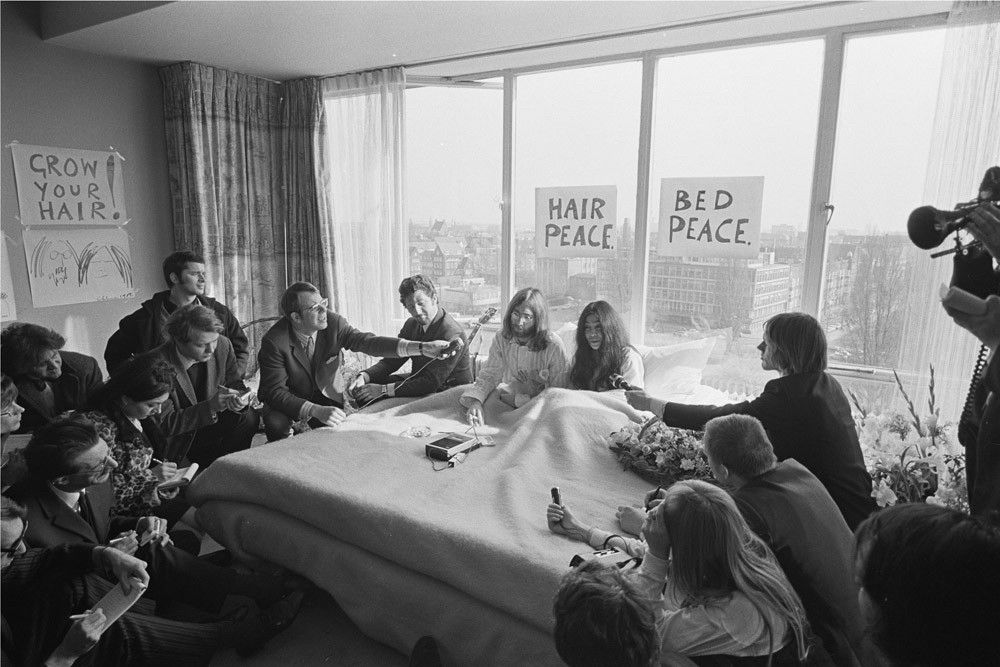 Press Conference in Pajamas – The Montreal Bed-In Begins

This black-and-white photograph captures a media-saturated moment during the opening day of John Lennon and Yoko Ono’s second “Bed-In for Peace,” launched on May 26, 1969, at the Queen Elizabeth Hotel in Montreal. The scene shows the couple seated in bed, smiling and dressed in white pajamas as they face a room packed with journalists, photographers, and documentarians. Around them, reporters sit cross-legged on the floor and lean forward eagerly with pens and notepads, microphones, and recording equipment, capturing every word and gesture. Behind the bed, large hand-lettered signs taped to the window panes declare their now-iconic slogans: “HAIR PEACE” and “BED PEACE.” The slogans were meant to subvert the expected modes of protest and instead present pacifism as both intimate and globally broadcastable. By inviting the media into their bedroom, Lennon and Ono inverted the private/public divide, turning the hotel suite into both a sanctuary and a stage. The Montreal Bed-In was not a spontaneous act but a carefully orchestrated performance of nonviolent activism, timed to attract maximum press coverage. It took place shortly after their wedding and was inspired by Gandhi’s methods of peaceful resistance. Unlike traditional protests marked by confrontation or marching, the Bed-In used the media spectacle — a tool Lennon and Ono understood deeply — to shift conversations toward peace and demilitarization. During this week-long event, the couple gave interviews to outlets around the world and ultimately recorded the anthem “Give Peace a Chance.” This photograph captures the very spirit of their protest: sincere idealism blended with theatrical showmanship, challenging viewers to rethink how peace could be enacted not just in the streets, but in the everyday act of being.
