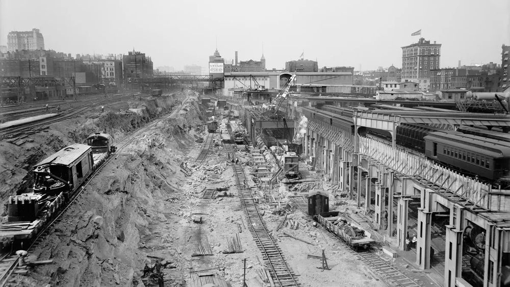 Excavation and Rail Infrastructure Development at Grand Central

This black-and-white photograph captures the massive excavation and infrastructure work undertaken during the construction of Grand Central Terminal. The image showcases a vast trench filled with railway tracks, partially constructed platforms, cranes, and workers engaging in complex engineering efforts to transform the site into one of the world’s most advanced transportation hubs. Before the terminal’s reconstruction, this area was home to steam locomotives, which created heavy pollution, noise, and congestion. The electrification of train lines required digging deep underground to accommodate a revolutionary two-level train system. This photograph reveals the significant demolition and earthmoving that took place to replace outdated facilities with a modern, efficient network. The image highlights the transition from steam power to electric railways, a major innovation of the time, allowing trains to operate within enclosed urban environments without producing smoke. By constructing tunnels and electrified rail lines, New York Central Railroad aimed to create a more organized and efficient railway system, enhancing commuter experience. The complexity of the construction process is evident in the unfinished tracks and exposed earth, demonstrating the immense scale of the undertaking. As part of this project, entire city blocks were excavated, requiring thousands of workers and years of labor. By the time Grand Central Terminal opened in 1913, it was not only the largest train station in the world but also a model for future transportation infrastructure projects. This image represents an era of grand engineering feats, where urban planning and technological advancements converged to redefine travel in the United States.