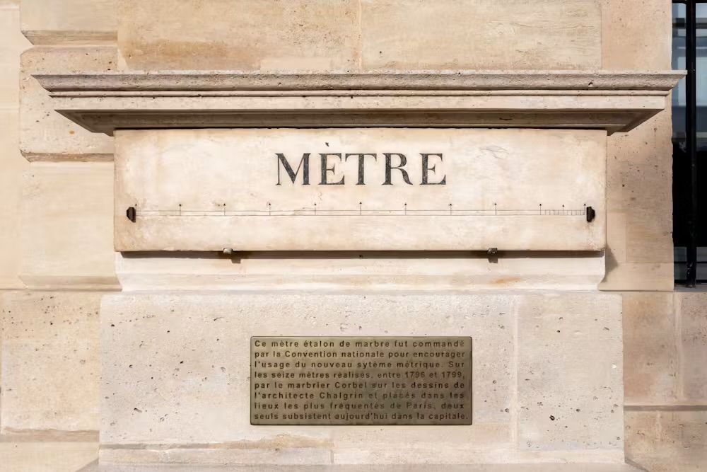 The Marble Metre – A Monument to Metric Standardization

This image showcases a historical marble metre standard affixed to a public wall in Paris, bearing the inscription “MÈTRE” in bold capital letters. Below the etched measurement line, a bronze plaque offers context in French: it explains that this standard metre was commissioned by the National Convention during the French Revolution to promote the use of the newly adopted metric system. Between 1796 and 1799, sixteen such metre standards were distributed across the most frequented locations in Paris. Only a couple remain today, making this one of the few surviving physical links to the dawn of global metric reform.

This physical artefact is a testament to the revolutionary ambition to rationalize and democratize measurements—a dramatic shift from the arbitrary, inconsistent systems previously used across regions and kingdoms. The metre was initially defined as one ten-millionth of the distance from the equator to the North Pole, a bold attempt to root human systems of measure in the natural world. The engraving above and below the line is both decorative and symbolic, intended to make the metre relatable and visible to the public at a time when education and civic unity were seen as revolutionary tools.

The installation of these standards in urban spaces allowed ordinary citizens to verify the length of goods in markets and trades, ensuring transparency and fairness. It reflects how the metric system was more than a scientific innovation—it was a sociopolitical tool for equity, knowledge, and national cohesion. The metre bar captured here is more than just a measure of length; it is a relic of enlightenment thinking, a marker of France’s pivotal role in shaping modern scientific standards, and a reminder that even the most ordinary-seeming measurements can hold profound historical significance.