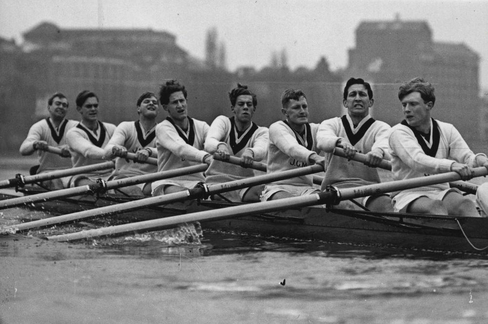 Oxford vs. Cambridge – Mid-20th Century Rivalry in Full Stroke

This powerful black-and-white image captures the Oxford rowing crew during a mid-20th-century edition of the Oxford-Cambridge Boat Race, likely in the 1950s or early 1960s. The rowers, clad in matching white sweaters with dark v-neck stripes, are caught in unison mid-stroke, their faces marked by focus and exertion. The boat cuts sharply through the water, oars perfectly aligned, reflecting the discipline and synchronicity required at the highest level of competitive rowing. The background shows a soft silhouette of London’s riverside architecture blurred by a foggy sky, situating the race in its historic setting along the Thames.

This era marked a golden period for the Boat Race in terms of both prestige and public fascination. Broadcast on radio and later television, the race attracted massive audiences across the UK and internationally. The 1950s and 60s saw an evolution in rowing technique and training regimens, driven in part by advances in sports science and growing university athletic programs. During this time, both Oxford and Cambridge began to more formally incorporate coaching expertise from former Olympians and national team rowers, pushing the level of competition to new heights.

The image exemplifies not just athletic prowess but also the camaraderie and pressure unique to team sports. Each athlete relies entirely on the rhythm, strength, and mental fortitude of those around them. Success is not individual—it is forged in absolute collective unity. This photo offers a compelling visual metaphor for cooperation, tradition, and the endurance of academic and sporting institutions.

The Boat Race during this period had become more than a university event—it was a cultural fixture. Political leaders, royals, and international dignitaries regularly attended. And yet, its essence remained unchanged: eight rowers and a coxswain, rowing not just for victory but for legacy.