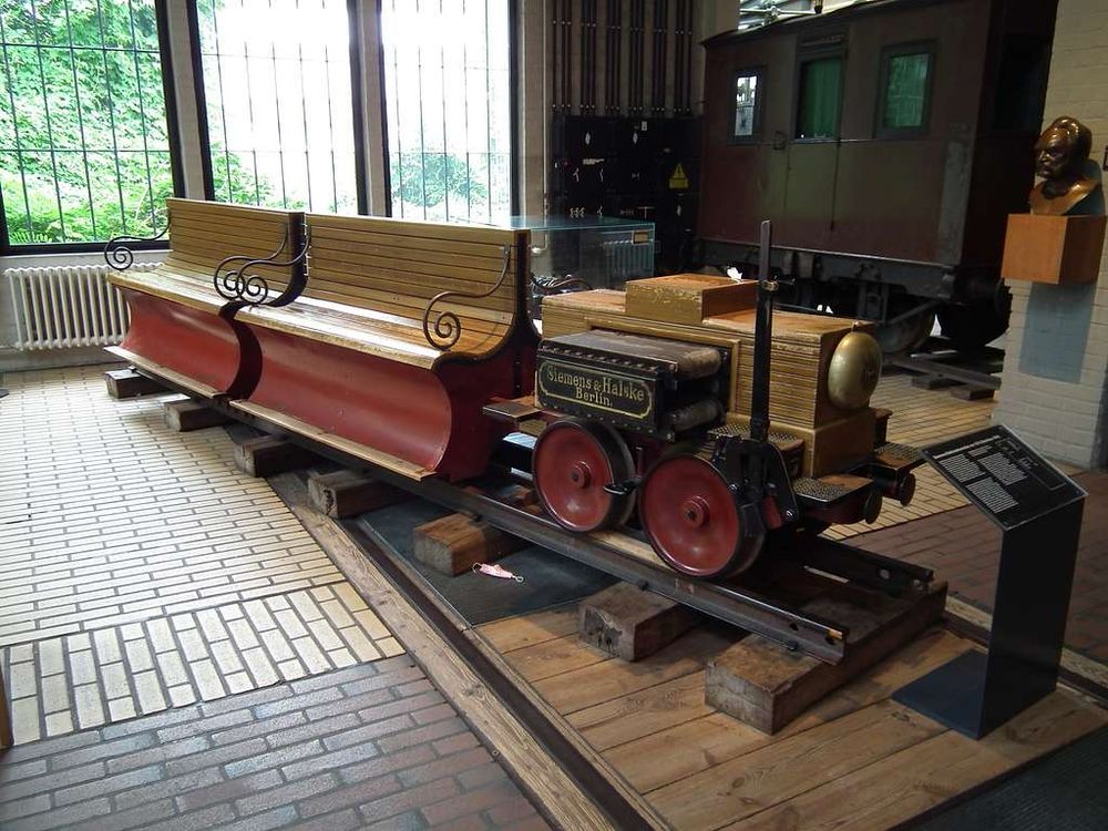 The World’s 1st Electric Locomotive Displayed at the German Museum of Technology

This image shows the preserved Siemens electric locomotive from 1879, housed at the German Museum of Technology in Berlin. The compact, wood-paneled locomotive, marked “Siemens & Halske Berlin,” sits on short rails within a polished exhibition room featuring historical rail artifacts and natural light filtering through iron-framed windows. With four small iron wheels and a brass cylinder casing, the locomotive’s design reflects both simplicity and mechanical ingenuity. Attached is an open-air bench car with finely finished wood seating and decorative wrought-iron side scrolls, designed to carry several passengers in rows. The locomotive and car rest on wooden supports to preserve their condition, and a black interpretive placard stands to the right, offering historical context for visitors

Unveiled on May 31, 1879, at the Berlin Industrial Exhibition, this locomotive was designed by German engineer Werner von Siemens. It ran on a 300-meter circular track and drew electricity from a third rail, not overhead wires—an experimental design choice that emphasized efficiency and novelty. Over 86,000 people rode the train during the exhibition, providing proof of concept for electric-powered transport. The motor used a direct current system powered by an external generator, setting a foundational standard for electric transit systems

This marked a pivotal transition away from steam propulsion, opening the door for electric trams, subways, and light rail networks that would revolutionize urban infrastructure worldwide. The locomotive didn’t just carry passengers—it carried society into a new era of motion, one defined by clean energy, reduced urban pollution, and the scalable electrification of transportation. As cities around the world now return to electric transit in pursuit of sustainable development, this artifact remains a powerful symbol of innovation’s potential to reshape daily life