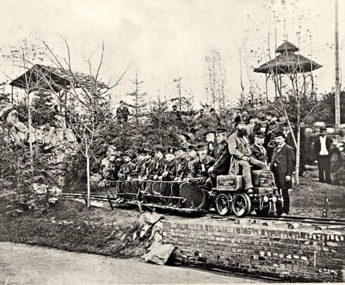 Siemens Electric Locomotive Demonstration at the 1879 Berlin Industrial Exposition

This photograph captures a pivotal moment in the history of transportation: the public demonstration of Werner von Siemens’ electric locomotive at the Berlin Industrial Exposition in 1879. Taken outdoors on a narrow-gauge test track constructed specifically for the event, the image shows the small electric locomotive—bearing the name “Siemens & Halske Berlin”—pulling a procession of passenger cars filled with dignitaries, engineers, and curious onlookers. The locomotive is driven by an operator seated upright at the front, hands on the controls, while Siemens himself is believed to be among the men overseeing the demonstration to the left. Visitors, mostly men in top hats and long coats, are packed closely on the open railcars, some appearing skeptical, others smiling in awe. In the background, trees and viewing platforms are filled with additional observers, and makeshift structures frame the pathway, suggesting the festive and experimental atmosphere of a world’s fair. The modest size of the locomotive stands in stark contrast to its groundbreaking implications. Powered by a 2-horsepower direct current motor supplied through a third rail—visible beneath the track—this prototype was capable of hauling up to 18 passengers at a time over a short loop. It was the first time in recorded history that electricity had been successfully used to propel a vehicle along a track with passengers aboard. The demonstration amazed thousands and sparked global interest in electrified rail systems. More than a novelty, this event planted the seeds of what would later become city streetcars, subways, and electric interurban trains. It marked the shift from coal-driven to current-driven transit, laying foundations for the modernization of global infrastructure. Siemens’ contribution, vividly brought to life in this image, redefined the boundaries of what was technologically possible in public transport