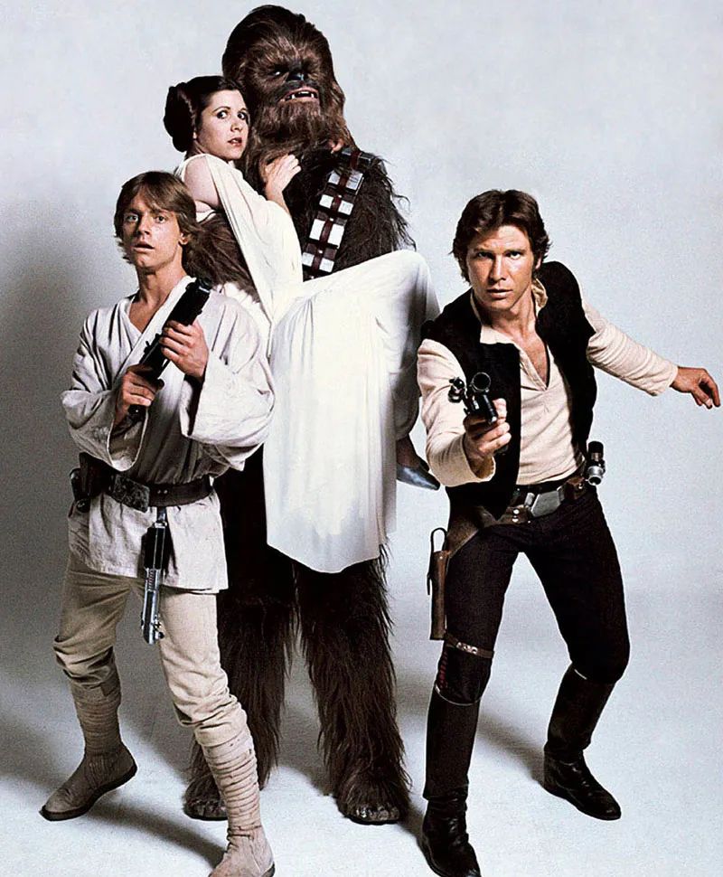 A Galactic Ensemble – The Iconic Cast of Star Wars (1977)

This promotional image showcases the principal heroes of Star Wars (1977), posed in character for a timeless ensemble portrait that would define the saga for generations. From left to right, Luke Skywalker (Mark Hamill), Princess Leia Organa (Carrie Fisher), Chewbacca (Peter Mayhew), and Han Solo (Harrison Ford) stand together in full costume, striking dynamic poses that capture the spirit and essence of the film’s mythic adventure. Hamill’s Luke stands at the ready with his blaster, symbolizing the reluctant farm boy turned Jedi-in-training. Fisher’s Leia, cradled in Chewbacca’s arm and wielding her own blaster, blends royal elegance with revolutionary defiance, embodying the feminist strength that set her apart from earlier Hollywood damsels. Chewbacca towers above the group in his full Wookiee costume—made of yak hair and mohair—exuding loyalty and power as Han Solo’s fiercely protective co-pilot. Ford’s Han stands confidently, blaster drawn, channeling the roguish charm and cynical bravado that made his smuggler-turned-hero one of cinema’s most beloved antiheroes.

Photographed against a clean studio backdrop, the image highlights each character’s design: Luke’s simple Tatooine tunic and boots, Leia’s flowing white gown with a retro-futuristic flair, Han’s vest and holster, and Chewbacca’s bandolier—each a deliberate fusion of science fiction and fantasy aesthetics. The actors’ expressions—some wary, some bold—mirror their characters’ roles in the galactic rebellion. This image was central to Star Wars marketing efforts, often reproduced in merchandise and posters. More than just a cast photo, it became an emblem of cinematic adventure, introducing a universe of archetypes reimagined through George Lucas’s futuristic mythos. It remains one of the most enduring images of 20th-century popular culture.