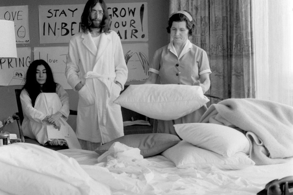 Morning Routine During the Montreal Bed-In (1969)

This black-and-white photograph captures a candid moment from John Lennon and Yoko Ono’s second “Bed-In for Peace,” held in May 1969 in Montreal. The image shows Lennon in a white bathrobe, standing beside Yoko Ono, who is seated cross-legged and barefoot in a chair, also dressed in white. A hotel maid stands at the center, fluffing pillows — a quiet symbol of domestic normalcy in an otherwise radical protest setting. Surrounding them are handmade signs hung on the wall that reinforce the playful yet pointed messaging of the protest: “STAY IN BED,” “GROW YOUR HAIR,” and “SPRING LOVE.” These slogans echoed the couple’s belief in passive resistance and symbolic acts over violence. The Montreal Bed-In, which began on May 26, followed a similar event in Amsterdam and was planned as a weeklong media-engaged protest against war, especially the Vietnam War. Instead of marching, Lennon and Ono invited the world’s press into their hotel room to discuss peace — turning what would otherwise be a private honeymoon into a global statement. This particular scene underscores the surreal blend of the mundane and the revolutionary: the couple framed themselves as ordinary people doing something extraordinary, elevating a daily ritual like staying in bed to an act of philosophical resistance. The presence of the maid also highlights the lived-in, ongoing nature of the protest — this wasn’t a staged photo op but a multi-day event that blurred the line between personal life and public activism. With every moment, including this one, the Bed-In invited onlookers to reconsider how peace could be performed — not with confrontation, but with presence and intentional absurdity.