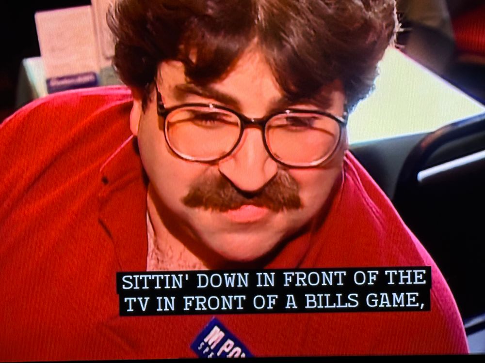 A picture of a man with a sweet mustache and ‘80s glasses sitting down at a restaurant table in the midst of talking about buffalo wings saying sittin down in front of the tv in front of the bills game. I believe it’s from an early Unwrapped episode.