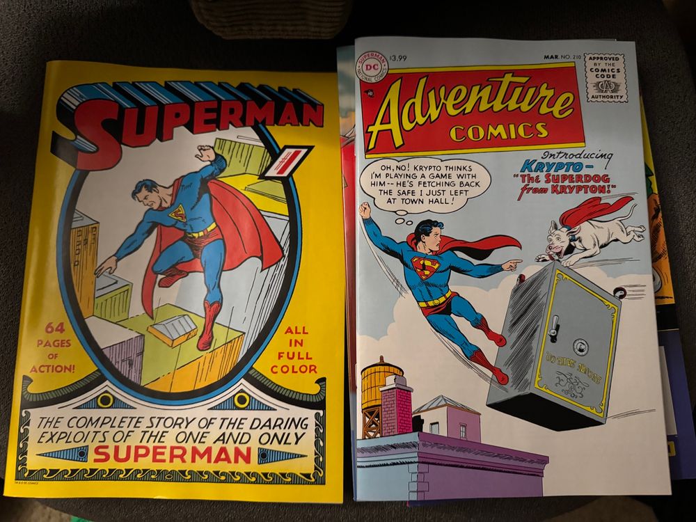 A picture of two comics from DC. Superman Number One, the complete story of the daring exploits of the one and only Superman. The cover is yellow and has Superman floating in the sky looking down at tall buildings. The second comic is Adventure Comics Number 210. It is the comic that introduces Krypto, the Superdog from Krypton. The cover features a flying Superman flying next to a floating safe that is being carried by Krypto. Superman is saying Oh no, Krypto thinks I’m playing a game with him. He’s fetching back the safe I just left at town hall. 
