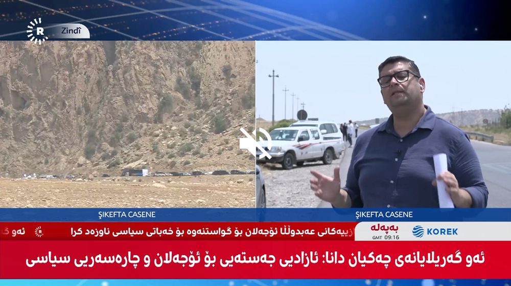Screenshot from a Rudaw split-screen of the PKK disarmament ceremony in the Kurdistan Region of Iraq. The left side is a static shot of the area outside Ranya, Sulaymaniyah, where dozens of cars and buses are parked up on the side of a mountain road. The ride side is the Rudaw correspondent on-site explaining the historic day. 