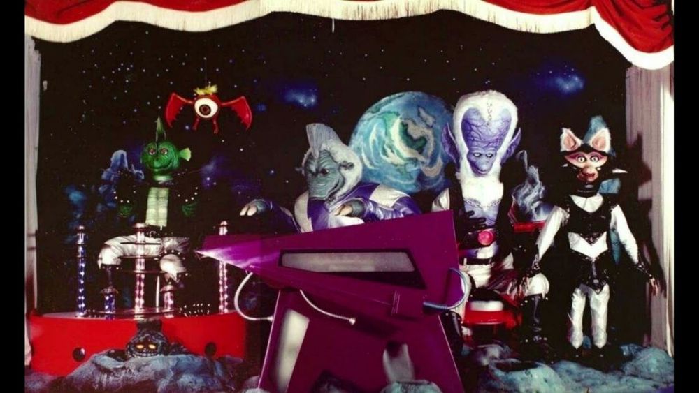 A photo of the Moonrockers on their stage. From left to right, the main animatronics are Ozone (a green lizard man playing the drums), Admiral Oort (an alien creature with white fur and grey-blue skin playing a keyboard), Quasar (a large-headed, blue skinned alien with white fur around his head and large blue ears), and Princess Haley (a cat or bat like alien with white fur, a silver and black outfit, and a cape). Above them is a small Moon Bat (essentially an eyeball with red wings) and below is a small rock creature in a moon crater.