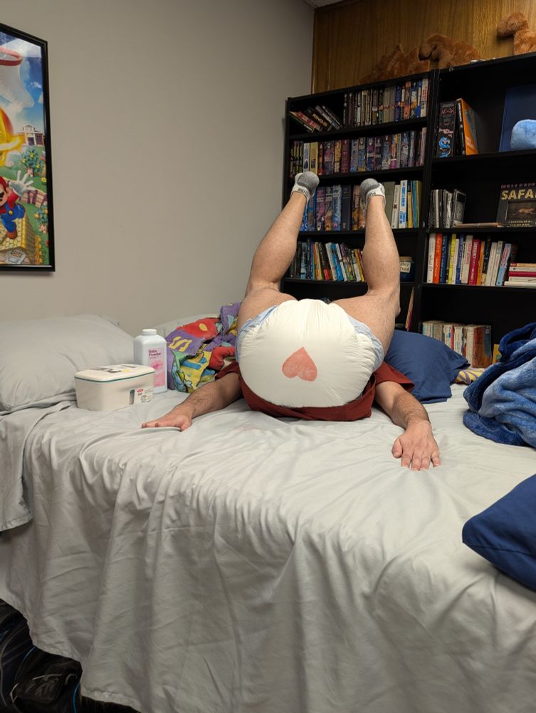 Adult guy laying on his back on a bed, legs lifted up showing off a round white diaper with a heart drawn on the butt
