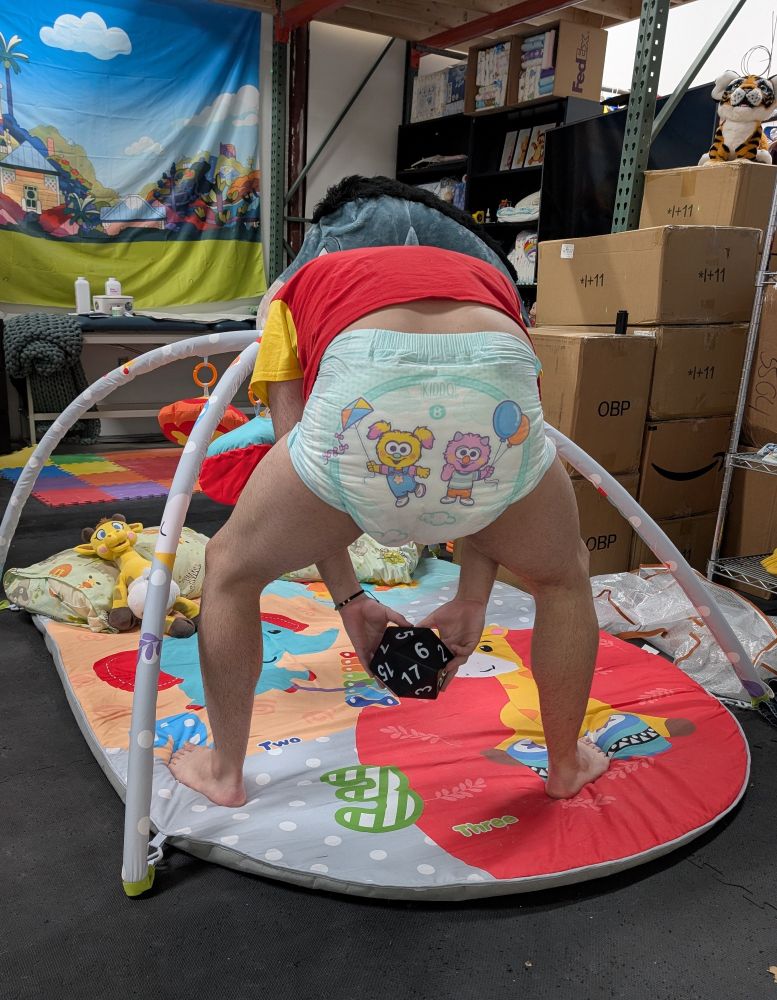 Adult male hunched forward facing away from camera. Wearing a cute adult printed diaper and no pants to cover it, standing at an adult-sized baby play gym as a cute colorful backdrop