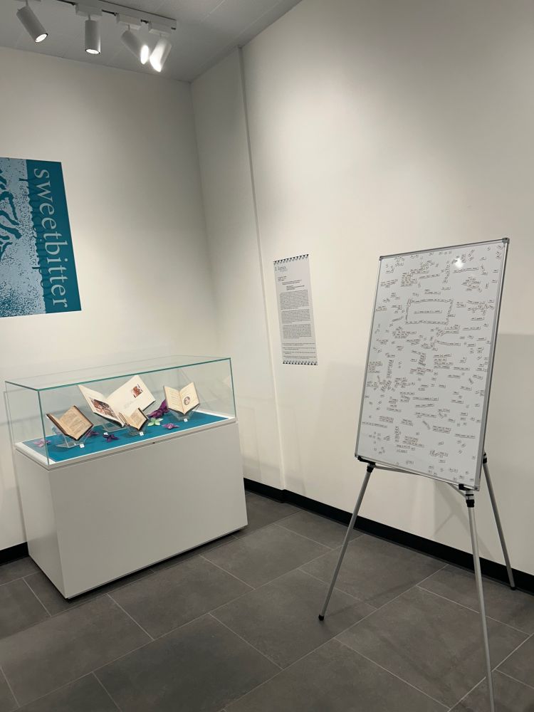 An easel with a whiteboard displaying magnetic strips with words on them, next to a display case with open books and a sign reading "sweetbitter."