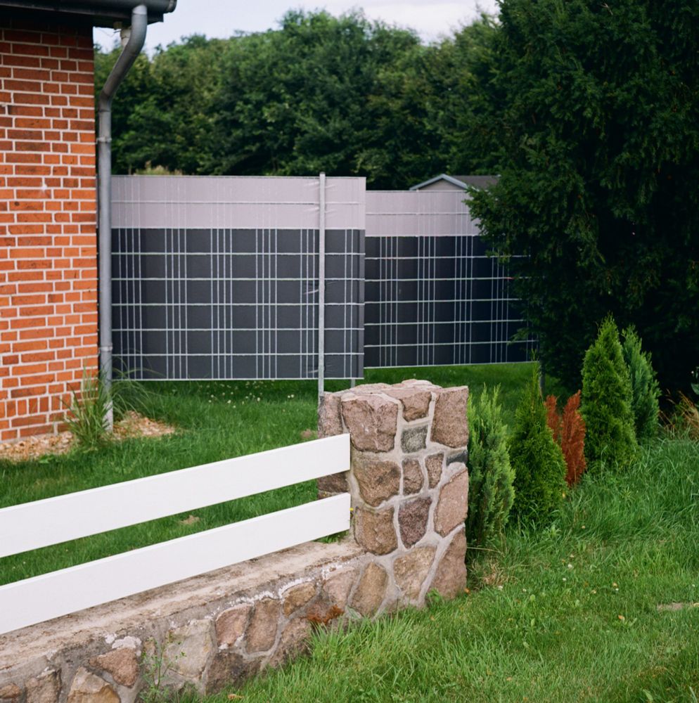 Auf der quadratischen Farbaufnahme ist ein weißer Holzzaun mit steinernem Unterbau und Pfosten aus Stein auf einem grünen Wiesenstück zu sehen. Links erkennt man eine geklinkerte Hauswand und dahinter einen weiteren, metallenen Zaun, der das Wiesenstück von einer Baumreihe trennt. 