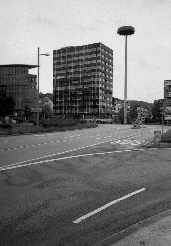 Ein etwa zehnstöckiges Hochhaus steht vor einer mehrspurigen Straße. Autos sind weit und breit nicht zu sehen, die Straße und die Fußgängerwegen sind menschenleer. Eine Straßenbeleuchtung erhebt sich rechts neben dem Haus, sie wirkt gigantisch groß. Auf der Straße sind die üblichen Fahrbahnmarkierungen zu sehen: Pfeile, Striche und das Wort Bus, das eine Busspur markiert. Die Szenerie wirkt trist