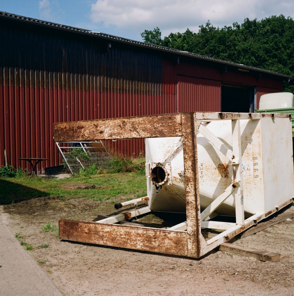 Auf dem quadratischen Farbbild ist ein umgekipptes Futtersilo mit rostigen Füßen zu sehen. Das Silo ragt schräg ins Bild hinein. Dahinter eistbein rostbraune Scheune zu sehen.