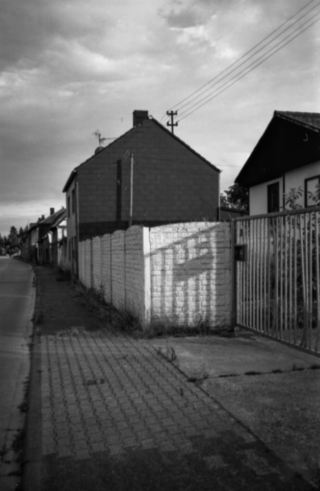 Blick in eine verlassene Straße, im Vordergrund eine weiße Mauer, auf die der Schatten eines Gottes fällt, dahinter ein dunkles Haus, vage erkennt man deb weiteren Verlauf der Straße, deren rechte Straßenhälfte man sieht. Dunkle Wolken lasten auf dem Bild.