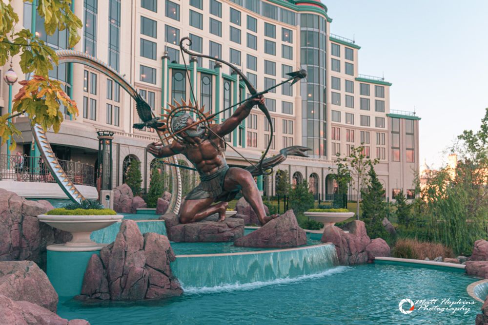 A statue with a bow and arrow stands over a waterfall with a hotel in the background.