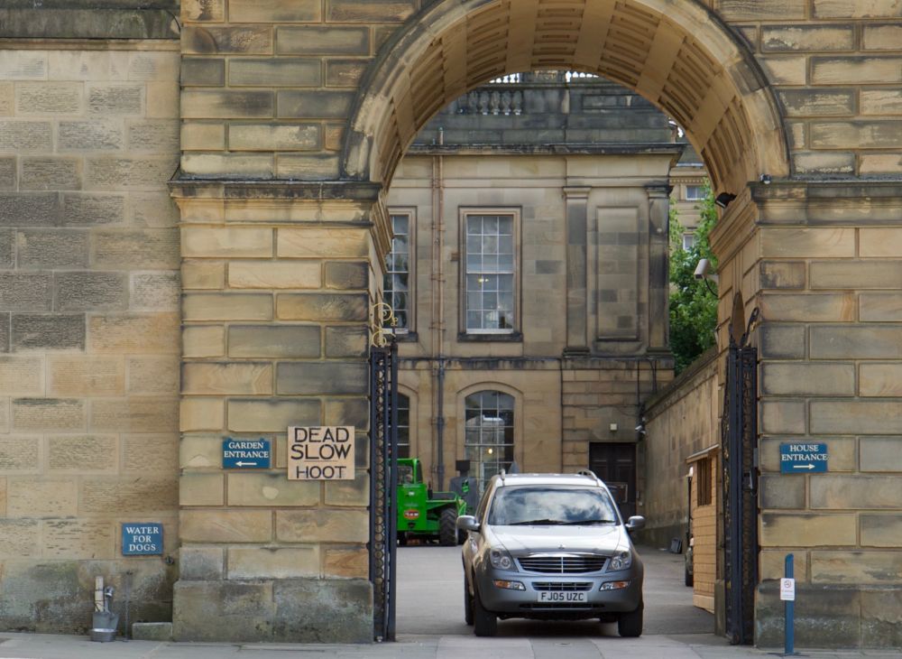 A car, facing us, is leaving the courtyard of a country manor through a large stone gateway. A sign on the left pillar of the gate says “DEAD SLOW / HOOT”