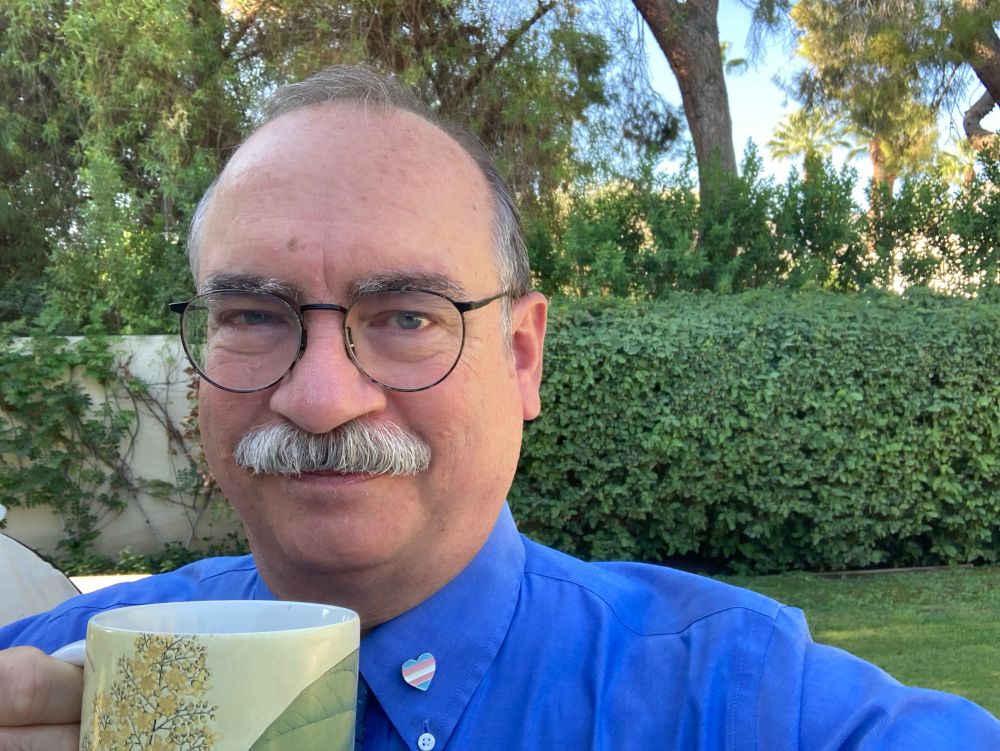 Everett Howe, a balding white man with a grey mustache and brown wireframe glasses, smiles while raising a yellow and green mug. He is wearing a blue dress shirt with a heart pin in trans pride colors on his collar, and he is standing in front of bushes and trees.