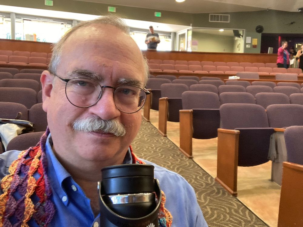 A selfie of a white man, balding, with a grey mustache and wire glasses, sitting in a pew in a mostly empty church sanctuary. He is holding a water bottle near his face. Around his shoulders is a colorful crocheted scarf. 