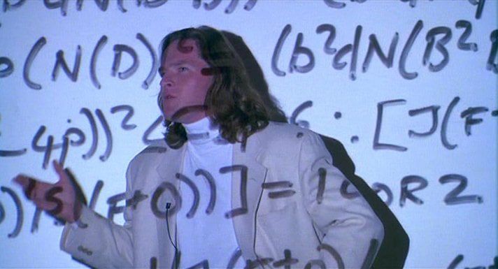 Donal Logue playing the mathematician Dr Gunter Janek in the movie Sneakers. He is wearing a cream sport coat over a white turtleneck, and he is standing in front of a white wall, lecturing. His handwritten slide transparencies are being projected on him and the wall; they are full of mathematical notation. 