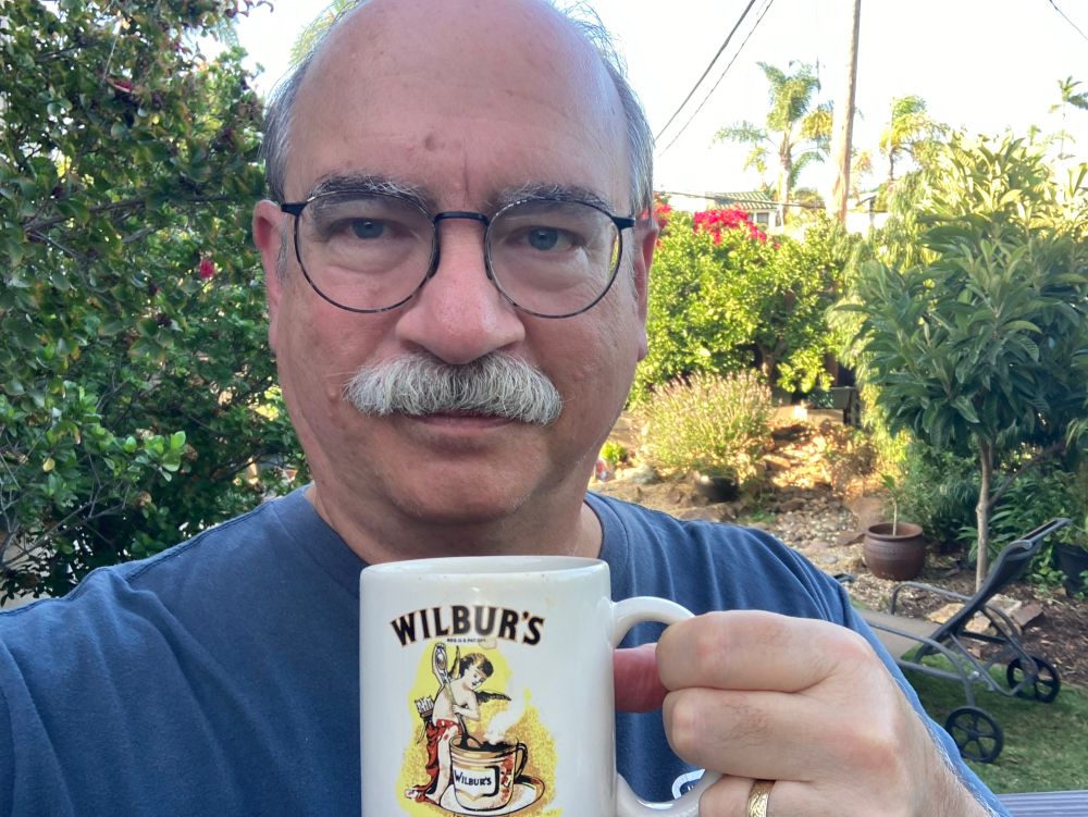 An older balding white man with glasses and a grey mustache, wearing a blue shirt and holding a coffee mug with an old-timey version of the Wilbur’s Cocoa logo on it. In the background are trees and bushes in a back yard. 