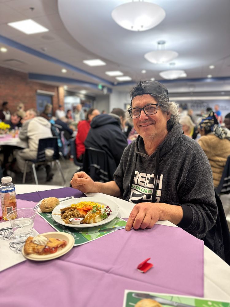 A client of The Ottawa Mission smiles with an Easter meal in front of him. In the background, you can see a dining room full of people eating Easter meals.