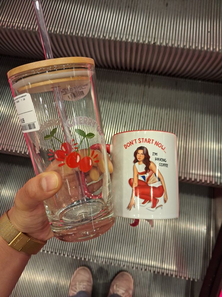 meine linke Hand hält einen Becher aus Glas mit einem Holzdeckel, der Becher hat einen Aufdruck von einem Paar Kirschen und der Aufschrift "Cherry good" und eine weiße Tasse auf der Dua Lipa zu sehen ist. dazu steht auf der Tasse "Don't start now - I'm having coffee".
