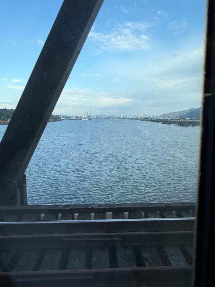 A photo of a wide river on a bright day, taken from inside of a train. Tall buildings are visible in the distance.