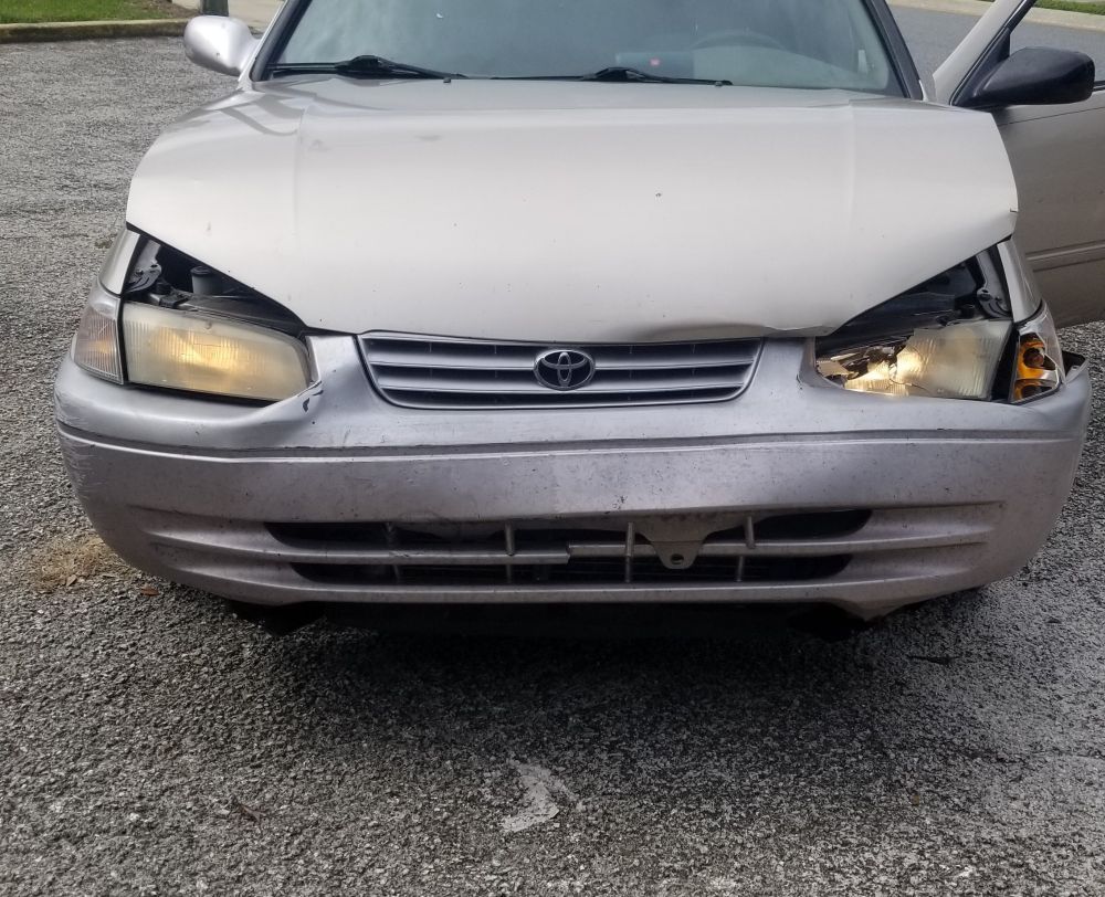 Front end of a wrecked toyota, driver side light busted, hood bowed and bumper smushed