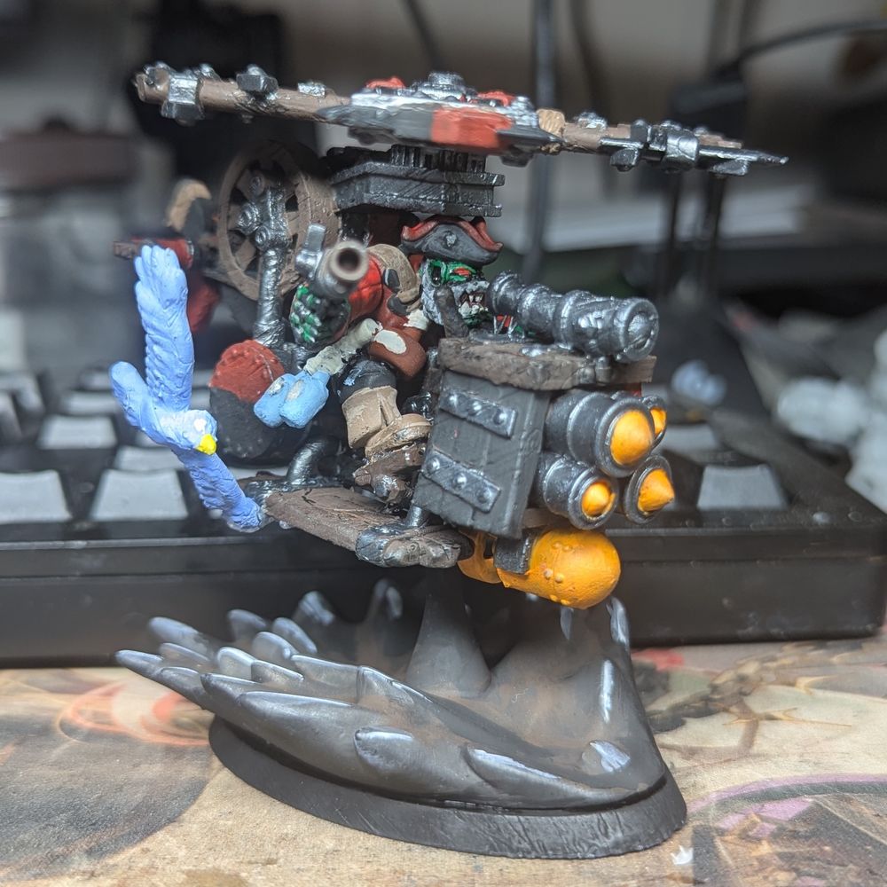 A crude gyrocopter armed with rockets on the front being piloted by an Ork in pirate attire holding a pistol. It's flying over oil and is accompanied by a blue sea bird flying with it.