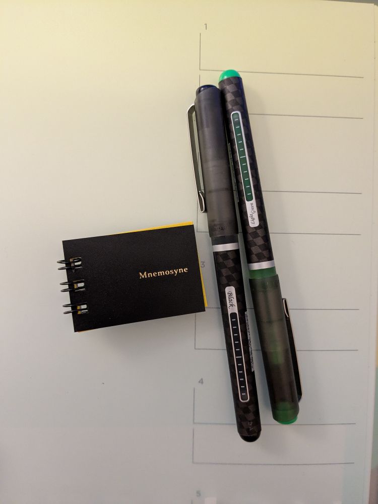 Two pens laying next to a thumb-sized black spiral notebook labelled "Mnemosyne".