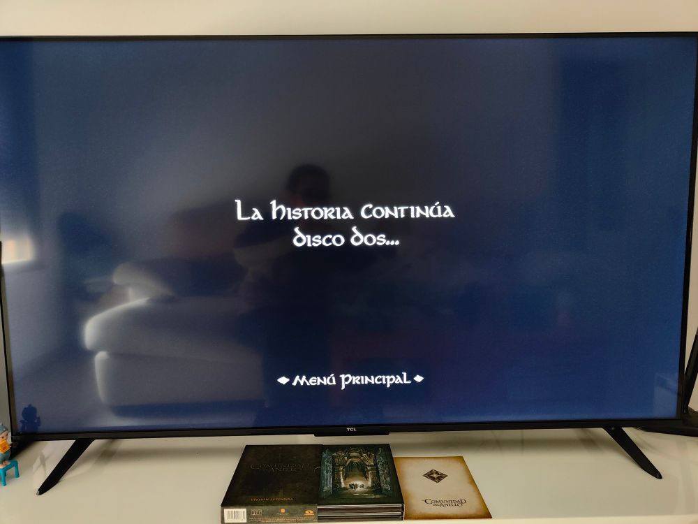 Pantalla del TV en negro con el texto "LA HISTORIA CONTINÚA DISCO DOS...", de El Señor de los Anillos Versión Extendida. Abajo, en el mueble del TV, la caja de La Comunidad del Anillo abierta, mostrando un arte conceptual de la Compañía del Anillo entrando a través de las puertas de Moria abiertas.