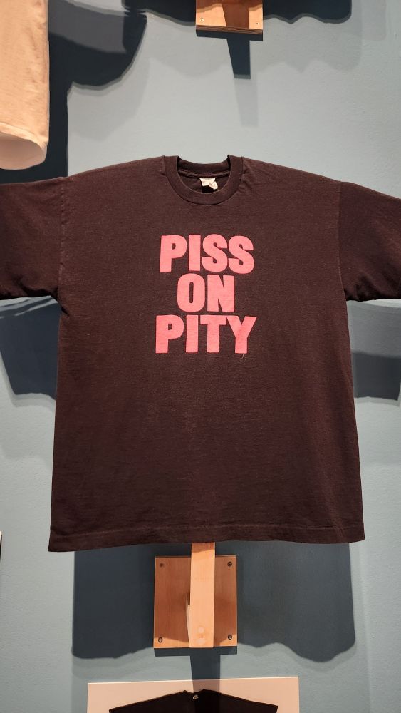 A black t shirt that says in bold pink writing Piss on Pity.