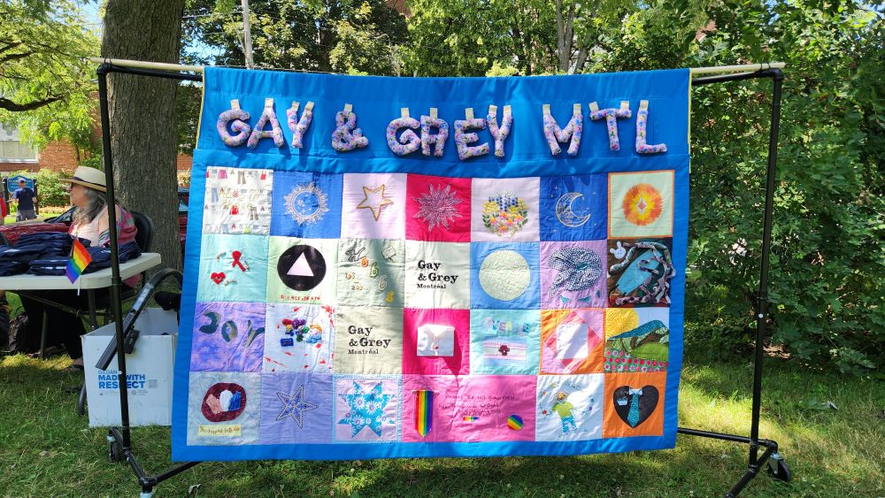 A photo of a quilt outside in a park hanging on a dowel. The framing of the quilt is royal blue. Large letters at the top in purple say Gay & Grey Montréal. 