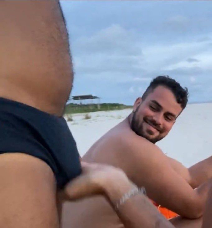 Uma mão feminina segurando o pacote de um cara usando sunga na praia enquanto outro cara olha um pouco atrás com um sorriso no rosto