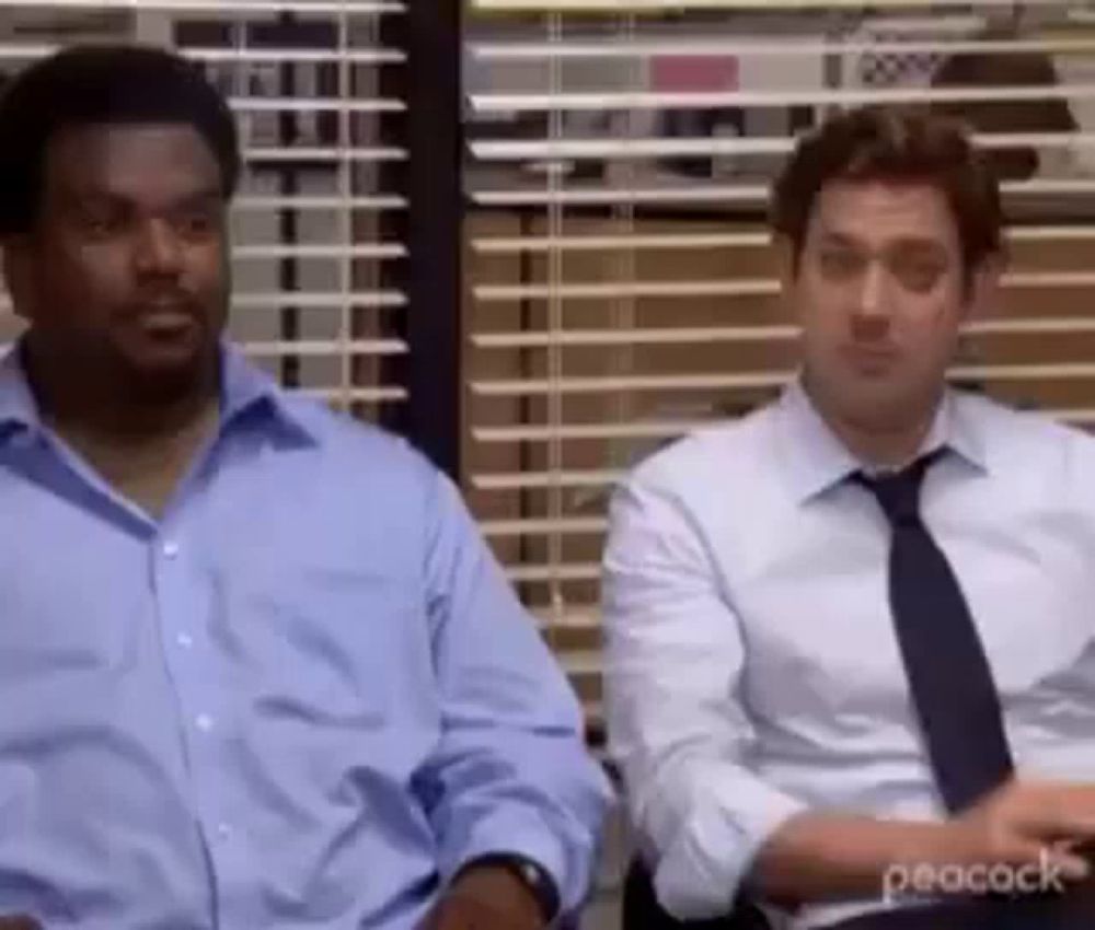 two men in suits and ties are sitting next to each other in front of a window in an office .