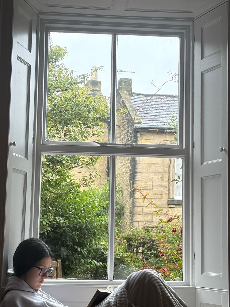 Teen girl reading a book on a rather lovely windiw seat. Leaves & bushes on the other side of the glass…