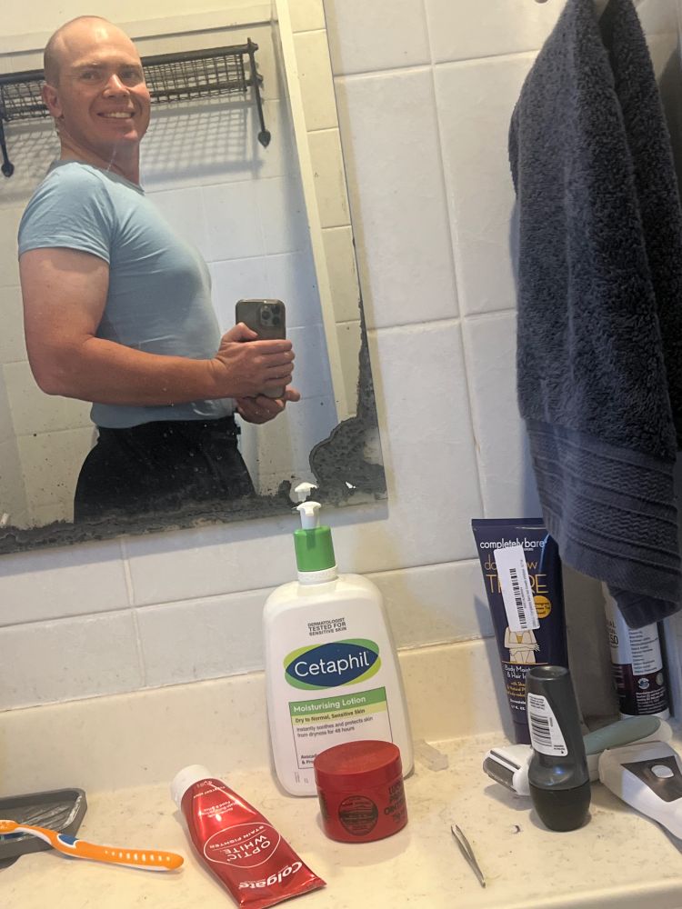A photo taken in a bathroom mirror of a moderately muscular, cis looking person with a balding head wearing clip on earrings, a skin tight light blue t-shirt and a black miniskirt. He has barely discernable breasts and is smiling.
