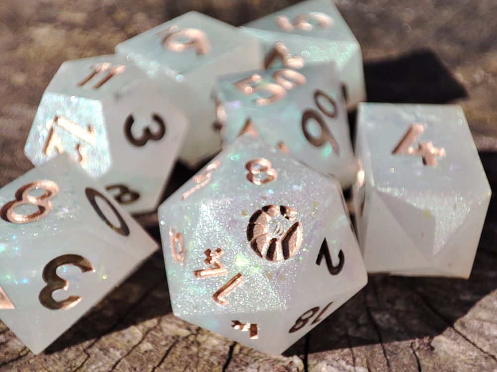 A 7 piece set of handmade resin polyhedral dice. They're designed after a frosted opal with glitter reflecting within a milky white base. They are inked in a rose gold/copper kind of colour, and the d20 is showcasing a shrimp logo on the 20 face. 
